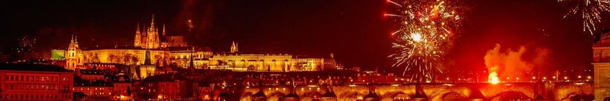 The city of Prague lit up by red fireworks, with the river flowing beneath the bridge