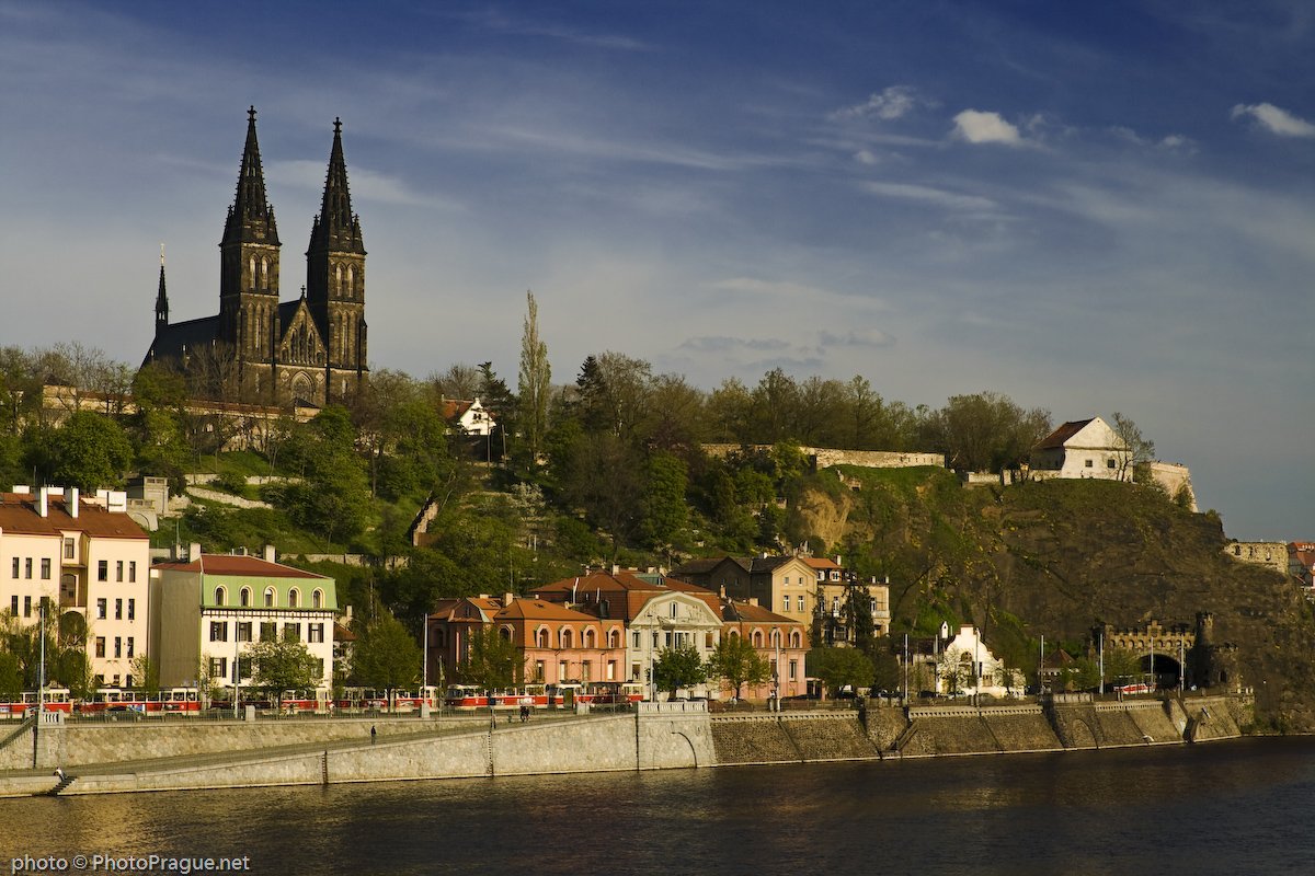 A fortified, verdant hill overlooks a river, with a church featuring two pointed spires.