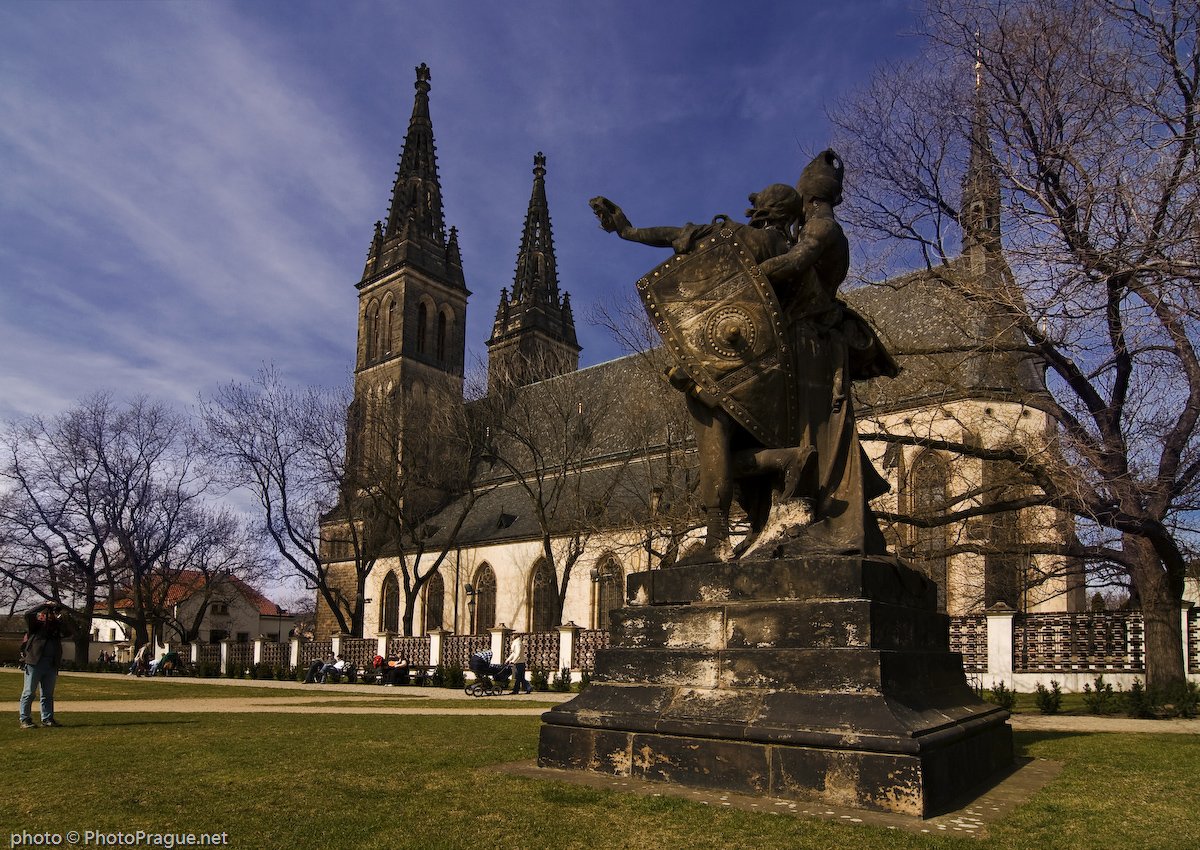 Stone statue of a warrior carrying a shield in the middle of a park, in front of a church with tall black spires