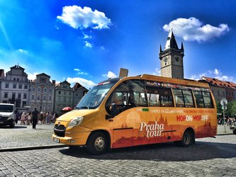 en:An orange and yellow tourist bus is parked in a sunny square, in front of old buildings and a tall clock tower. ;fr:Un bus touristique orange et jaune est stationné sur une place ensoleillée, devant des bâtiments anciens et une haute tour d'horloge