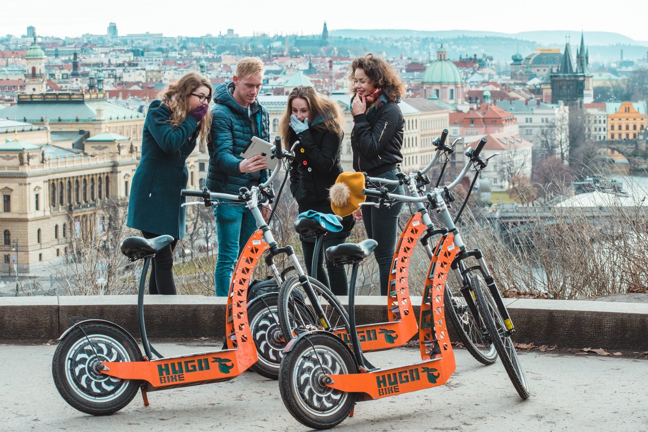 4 people looking at a tablet with surprised looks,standing in front of 3 electric scooters,with a view of the city