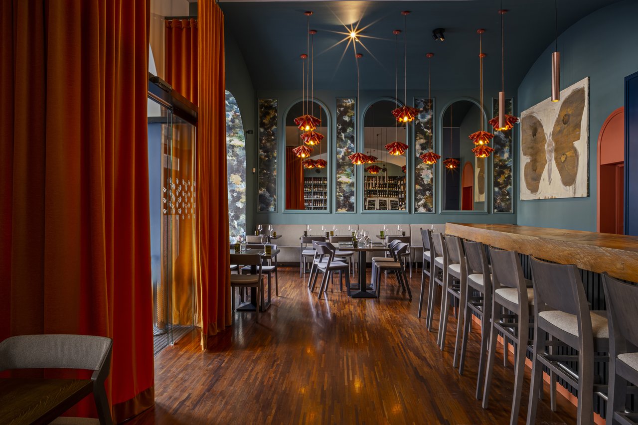 Inside the restaurant, there is a long table on the right, a painting of a butterfly on the wall, and red curtains on the left