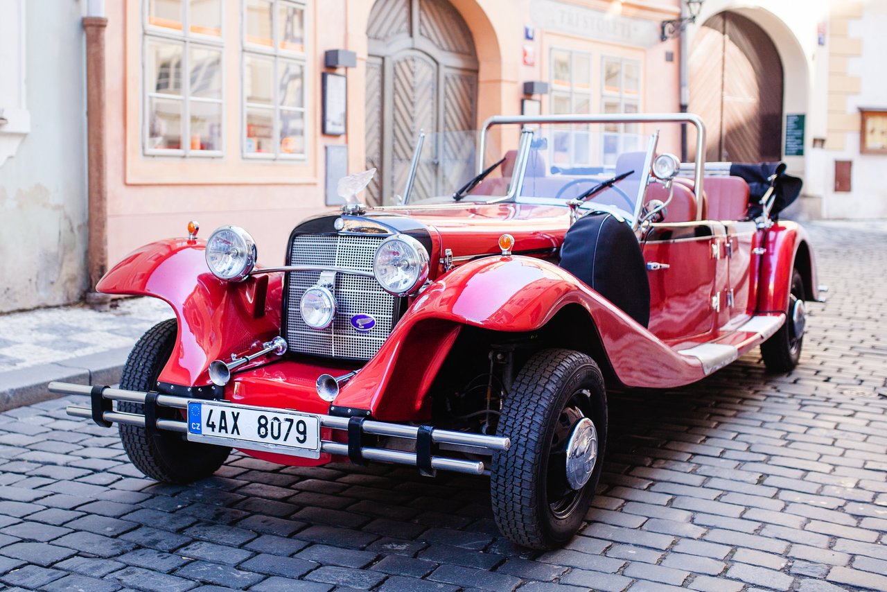 A bright red vintage convertible is parked on a cobblestone street in front of historic buildings in Prague.