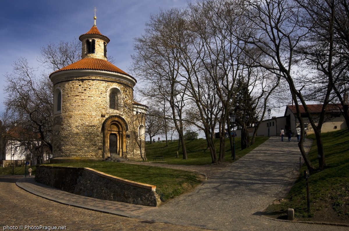 A small, round church made of light-colored stone with an orange tiled roof stands beside a paved path surrounded by leafless trees.